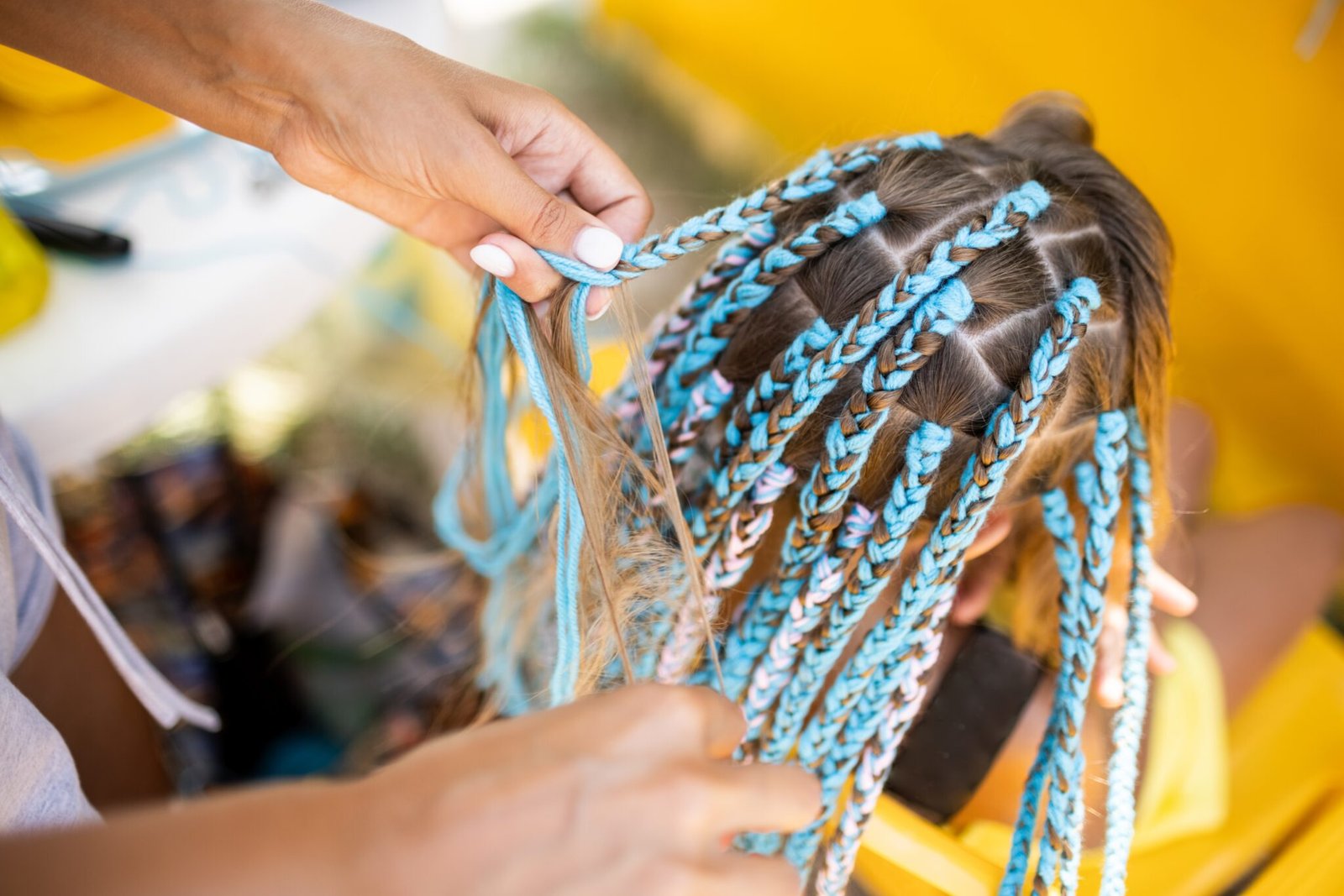 A little waiting tanned girl in a bright yellow summer suit, weaves colored pink-blue African braids in her dark wet thick hair on a hot sunny sea day