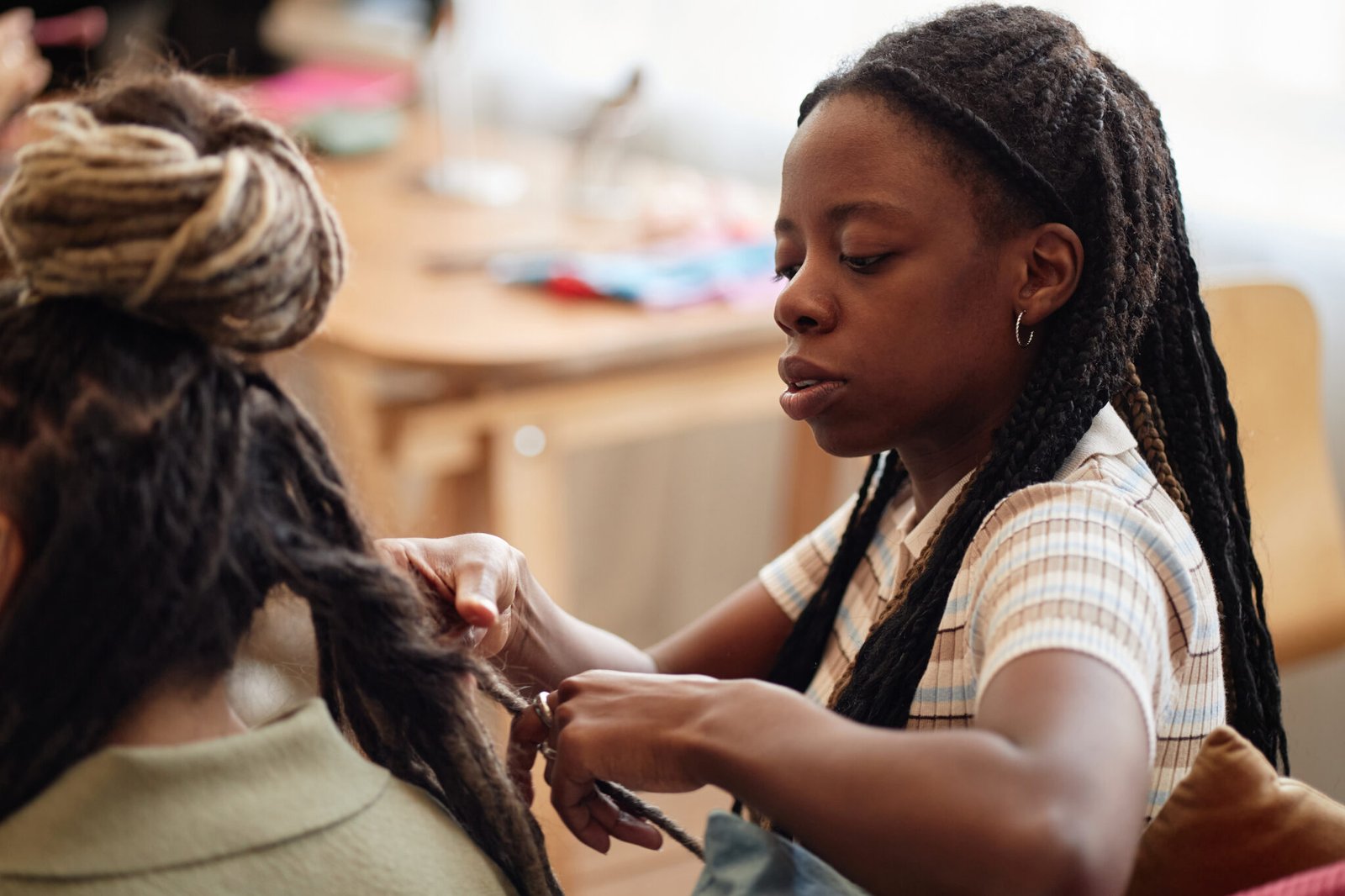 Black young adult woman braiding hair of girlfriend in indoor setting, focusing on hands and hair styling process, both women having long braided hairstyles, natural light background